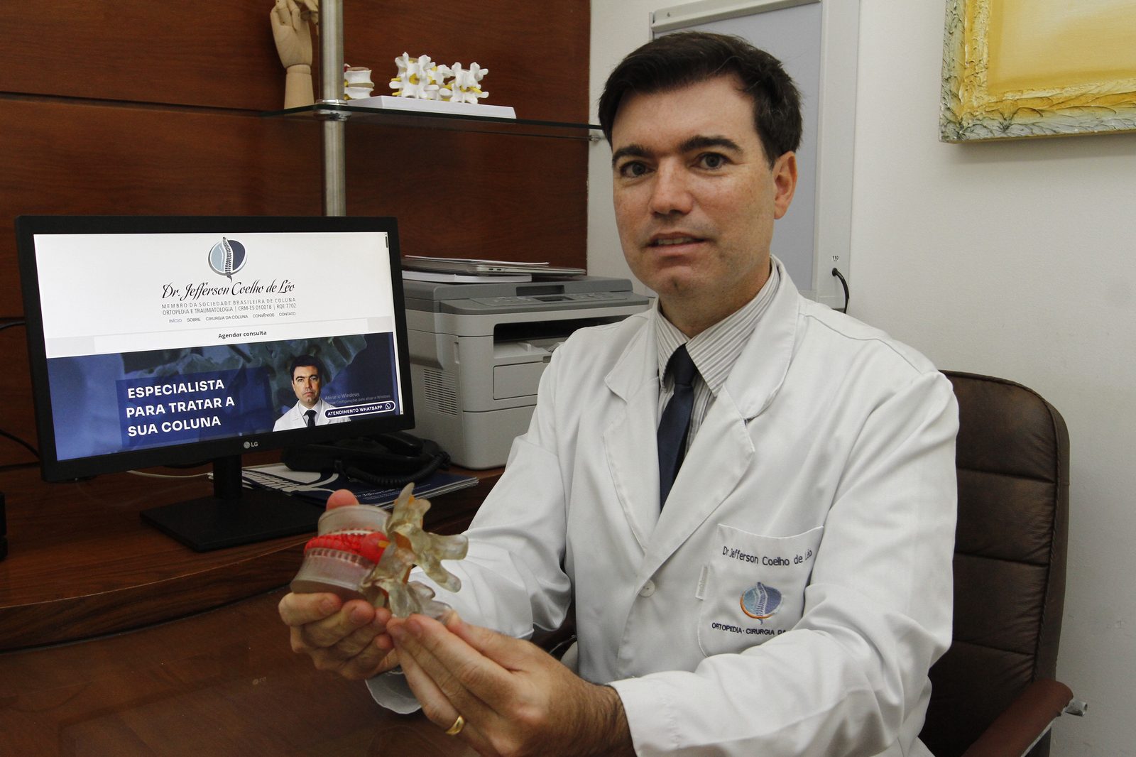 Dr. Jefferson Coelho de Léo em consultório, com modelo anatômico de vértebras, em Vitória-ES